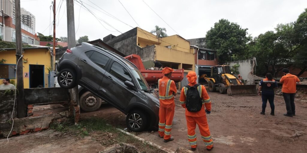Agentes de colete laranja ao lado de carro arrastado pela enxurrada numa rua com lama