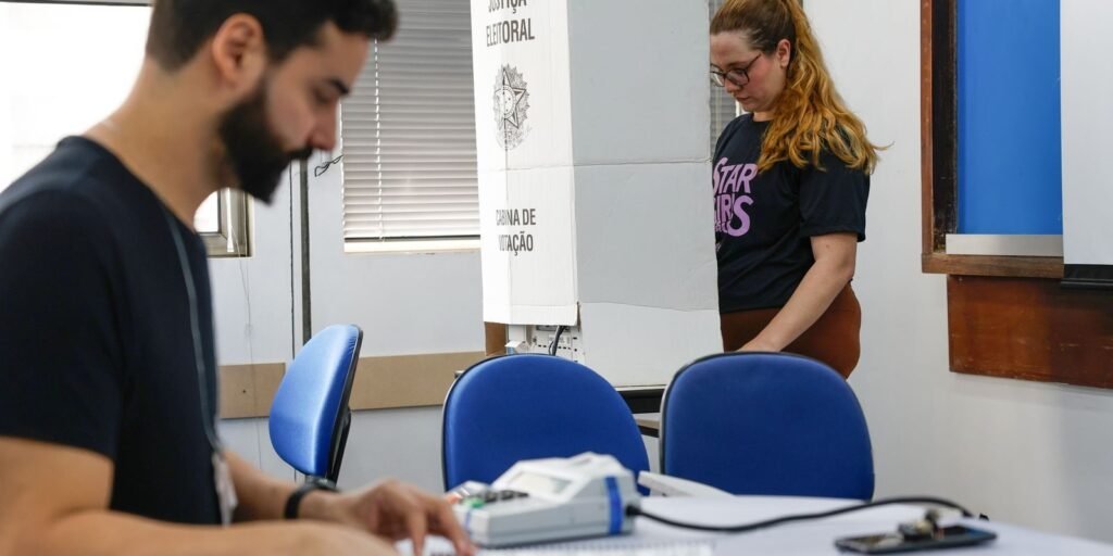 Mesário e eleitora em seção eleitoral com cabine de votação ao fundo