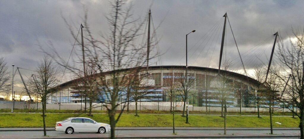 Estádio Etihad visto de longe, atrás de árvores sem folhas