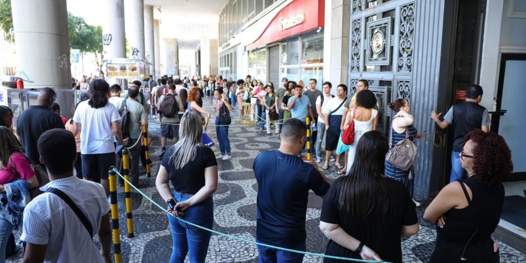 Fila de candidatos na calçada diante do portão de um prédio de prova