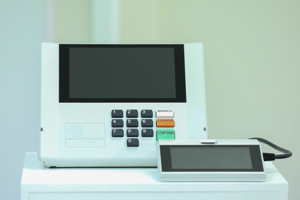 Urna eletrônica brasileira com teclado numérico e teclas Branco, Corrige e Confirma, ao lado do terminal do mesário